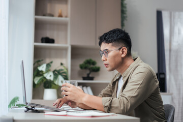 Fototapeta premium Asian man at a home office looks at his laptop while holding a coffee glass. Open notebook and pen on the desk, with soft daylight and green plants creating a neat, modern space for remote work.