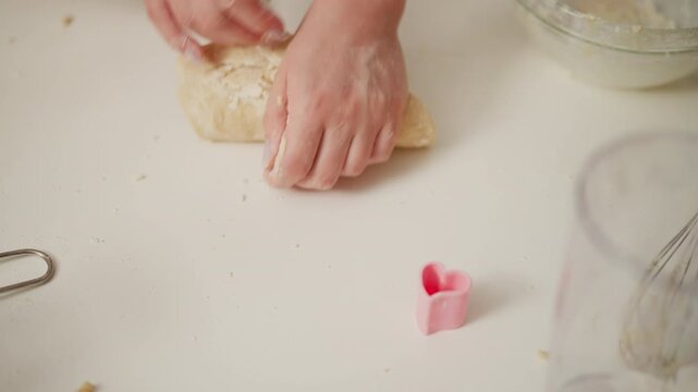 Hands moulding soft dough on floured table during baking process with person taking away roller pin, showing preparation stage with baking tools and pink heart shaped cookie cutter nearby