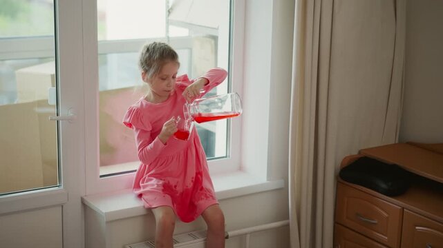 Niece seated by transparent window in daylight concentrating to pour red juice from glass pitcher into clear cup, small hands gripping handle, careful positioning to prevent spill on pink dress