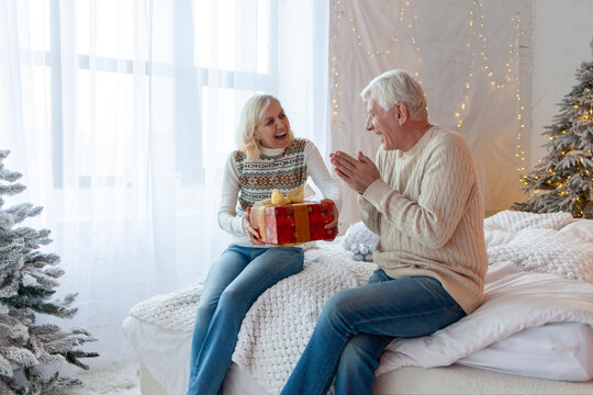 elderly couple of seniors in sweaters celebrate new year at home and exchange gifts, grandmother gives gift to husband and wishes merry christmas - Powered by Adobe