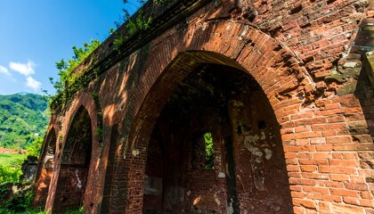Fototapeta premium A weathered brick archway structure, sun-drenched, displays remnants of a once grand edifice, surrounded by lush greenery and a distant mountain range.