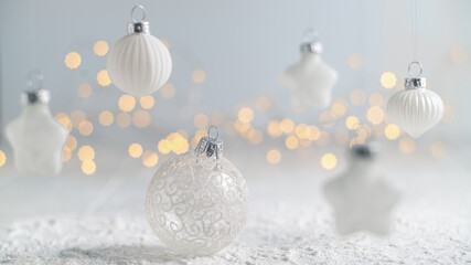 White christmas ornaments of various shapes hanging against a light background with soft, warm bokeh from sparkling lights, evoking a serene and festive holiday mood