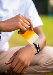 Black african man wearing white shirt, khaki trousers, wrist watch squating whiles holding yellow card outside .