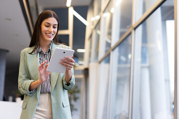 Businesswoman smiling while walking through a modern office hallway, using a digital tablet to stay...