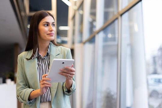 Businesswoman holding digital tablet looking out large office window contemplating future business vision