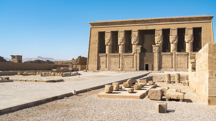 Facade of the famous Dendera temple, Egypt, dedicated to Hator goddess, with diagonal entrance path with ruins at both sides. Blue sky on the background.