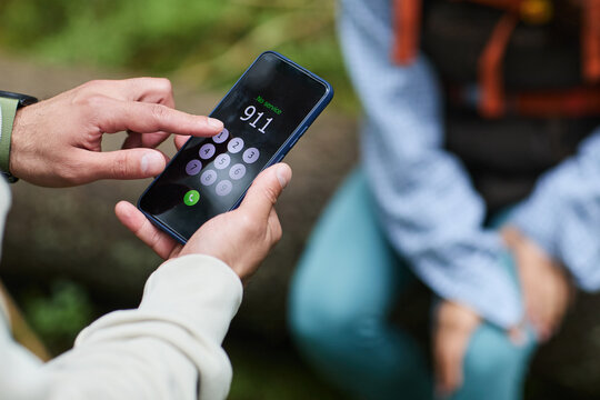 Young adult Caucasian man holding smartphone dialing emergency number 911 outdoors while sitting with another , focusing on hand and phone screen, partial body visible