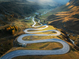 Aerial view of winding mountain road, river in alpine valley at sunset in autumn. Top drone view of road in Swiss mountain pass, yellow grass, trees in fall. Colorful landscape. Alps in Switzerland