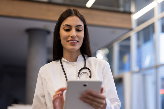 Smiling female doctor wearing lab coat and stethoscope using digital tablet in hospital corridor