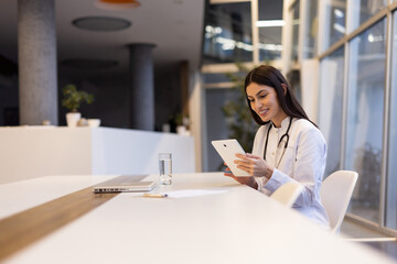 Smiling female doctor using digital tablet while sitting at desk in modern hospital office, healthcare and technology concept