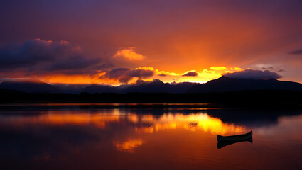 Fototapeta premium A lone canoe drifts peacefully on a calm lake as the vibrant orange and purple hues of a dramatic sunset paint the sky, reflecting beautifully on the waters surface