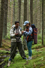 Fototapeta premium Young adult Caucasian man and young adult Asian woman standing in forest holding camping gear, smiling and looking at smartphone together, surrounded by tall trees and moss