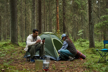 Fototapeta premium Caucasian young adult man sitting on folding chair using smartphone, while Asian young adult woman kneeling near tent preparing camping gear in forest with portable stove visible
