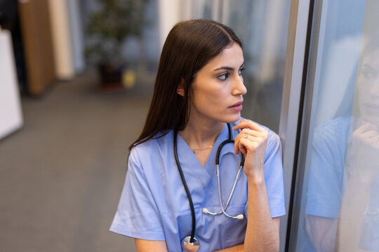 Pensive female doctor wearing blue scrubs and stethoscope looking thoughtfully out window in hospital corridor