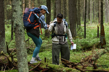Young adult Caucasian man helping young adult Caucasian woman cross fallen tree while hiking in forest, both carrying backpacks and navigating woodland terrain with map in hand