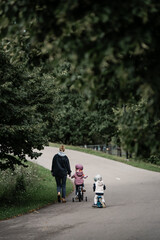 Parent walking with children on bicycle and scooter along park pathway, peaceful family outdoor activity in natural environment showing companionship, leisure and childhood mobility moment