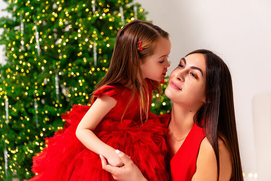Cute little daughter in red fluffy dress kissing her mother while they are sitting near Christmas tree, celebrating holidays. Parenthood and happy family concept. Merry Christmas and Happy New year