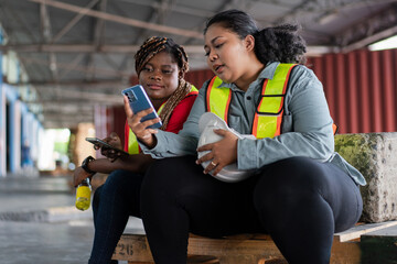 Two female African workers take a break, sitting and watching on mobile phone. chatting, sending message and browsing the internet at industrial factory
