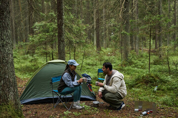 Young adult Caucasian woman and young adult Caucasian man sitting in forest near tent sharing hot drinks, camping equipment and supplies scattered on ground, surrounded by trees