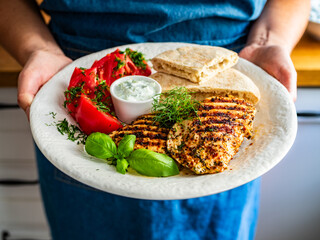 Woman holding plate with grilled chicken breast, tomatoes, pita bread and tzatziki sauce, close-up