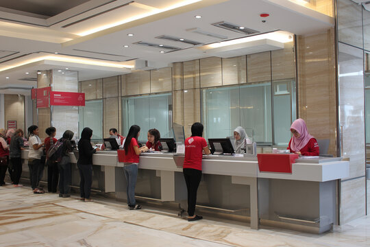Two bank tellers working side by side, the atmosphere is busy with a queue of customers.