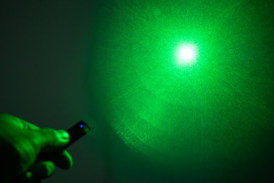 A person's hand holding a remote with a glowing blue light and a green laser beam pointing upwards in a hazy atmosphere.