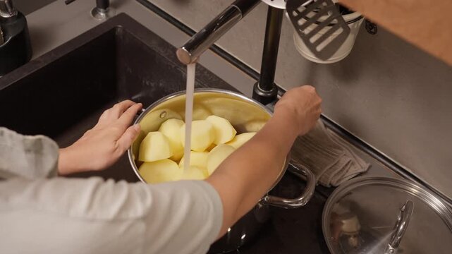 A housewife pours fresh tap water from a kitchen faucet into a large pot of halved potatoes. A simple, intermediate step in a recipe for preparing a homemade dinner before boiling.