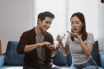 Young man taking care of his sick girlfriend, giving her medicine and a glass of water while sitting on the sofa at home