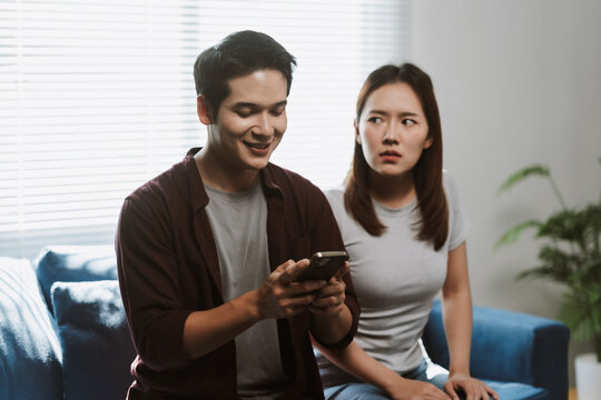 Young Asian couple experiencing relationship issues due to a smartphone, navigating emotions of jealousy, mistrust, and frustration while sitting on the sofa