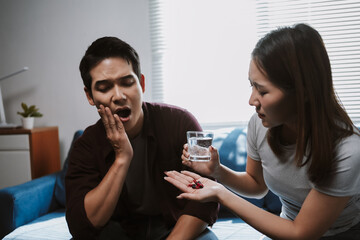 Asian couple in living room, girlfriend is taking care of her boyfriend who is suffering from toothache, she is giving him medicine and a glass of water