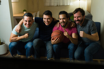 Happy Latin male friends cheering while watching basketball game on tv at home