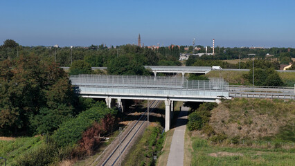 Obraz premium Modern infrastructure project under construction featuring concrete overpass and railway tracks surrounded by lush green trees and distant urban landscape under clear blue sky