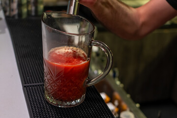 A bartender has finished pouring a red liquid into a glass pitcher, creating a vibrant red cocktail Sangrita with a frothy top, ready to be served