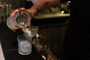 A bartender pours a golden cocktail from mixing glass with strainer into a glass filled with large...