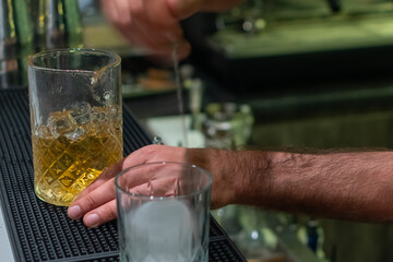 A bartender stirs a golden cocktail in mixing glass filled with ice, while another glass with large ice cube sits in foreground