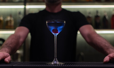 Vibrant blue cocktail Aviation in stemmed coupe glass close up against background with a bartender and bar shelves with bottles