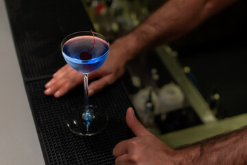 A vibrant blue cocktail garnished with dark cherry in stemmed coupe glass on black bar mat with a bartender's hands nearby