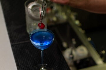 A bartender carefully using tongs to place a bright red cherry into vibrant blue cocktail in stemmed coupe glass