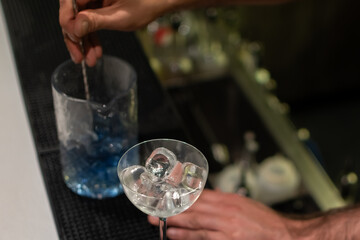 Bartender using long spoon to stir martini glass filled with ice with tall glass containing blue liquid in background