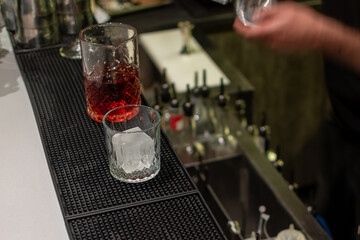 A glass filled with deep red cocktail and nearby glass with single large ice cube rest on bar mat, with bartender's hand in background