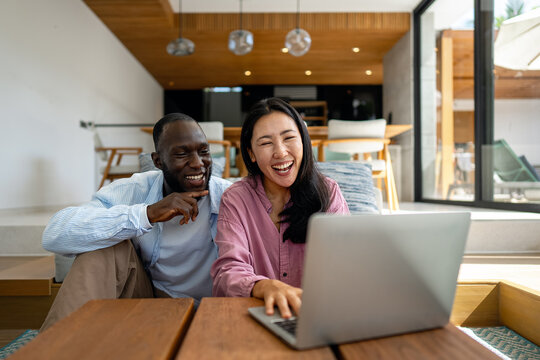 Young happy multiethnic couple having fun while using laptop at home. Happy couple discussing future