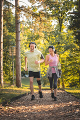 Fototapeta premium Happy diverse middle-aged couple jogging in forest, listening to music on headphones, enjoying outdoor exercise together, smiling and staying healthy in nature at sunset
