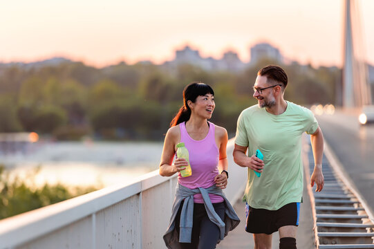 Smiling Asian woman and Caucasian man jogging together on bridge at sunset, enjoying outdoor fitness, diversity, and healthy active lifestyle