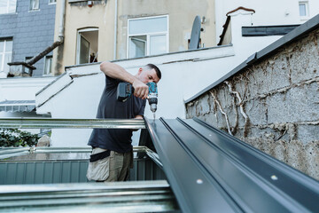 One mature handyman with a drill installs roofing sheet on a metal shed. © Nataliya