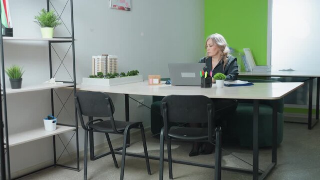 Sales woman seated at office desk working on laptop pauses to take latte before continuing, surrounded by model structure, organized desk items