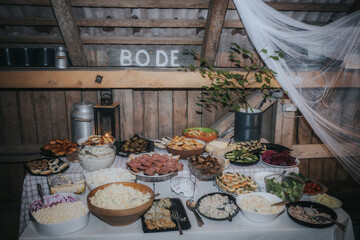 A rustic buffet table is filled with assorted traditional dishes, fresh salads, meats, and pastries, set in a cozy barn atmosphere.