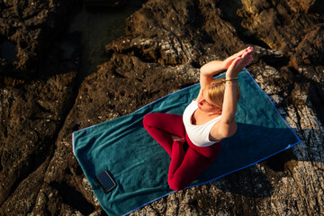 Practicing yoga on a towel along a rocky coast at sunset, a young woman embraces peace and mindfulness, surrounded by the serene beauty of nature's evening glow