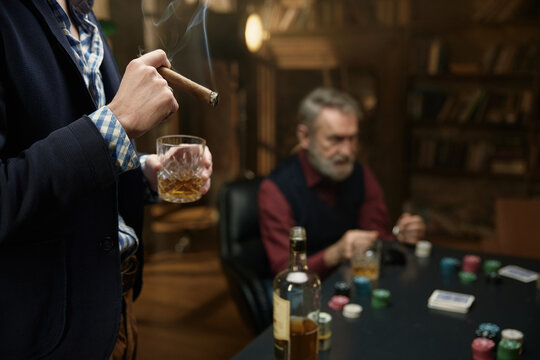 Man holding cigar and whiskey with his game partner on background while playing poker