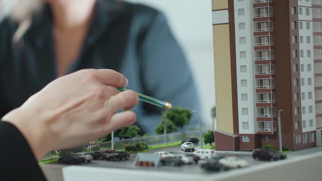close up hand view of real estate agent holding pencil explaining project to client using architectural building miniature surrounded by small cars trees and road layout in office setting