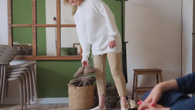 Visitor reaches into basket to pick crocs while psychologist in blue attire sits on sofa nearby, soft natural light fills room with mirror reflection, and casual welcoming therapy setting